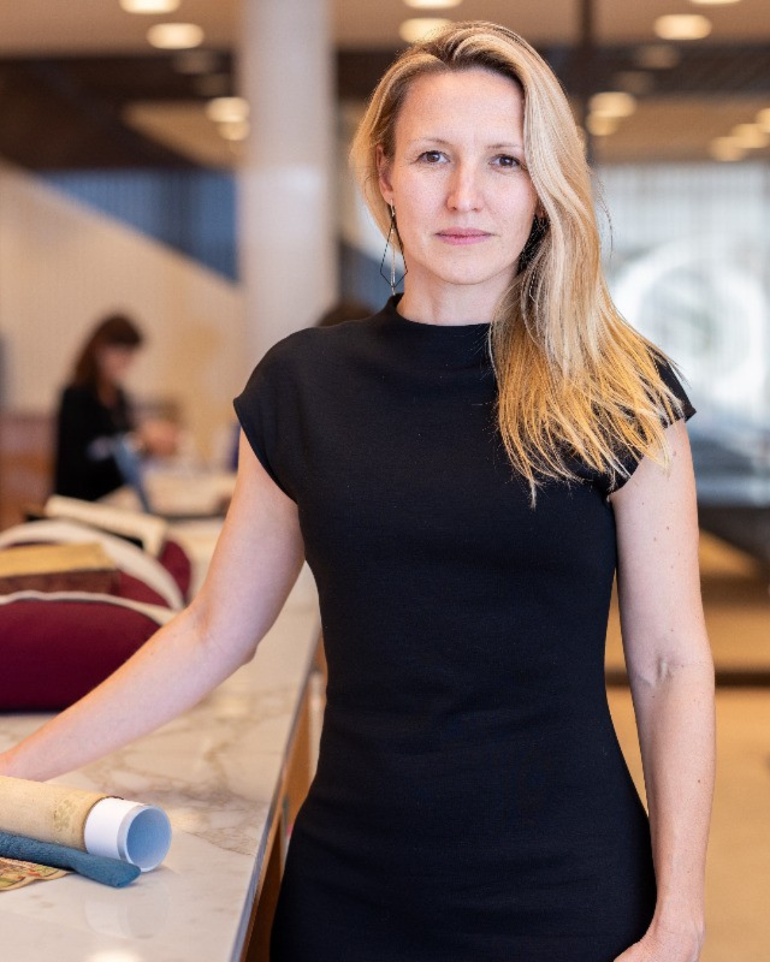 Photo of Agnieszka Rec, wearing a black dress stands by a marbled countertop with rolled papers.