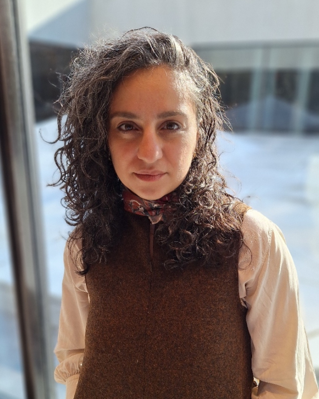 Photo of Michelle Al-Ferzly, wearing a brown vest and a scarf and standing indoors near large windows.