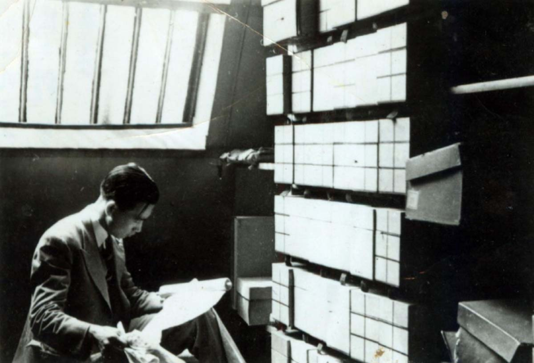 A person in a suit examines a document in a room with shelves of boxes and a large window.
