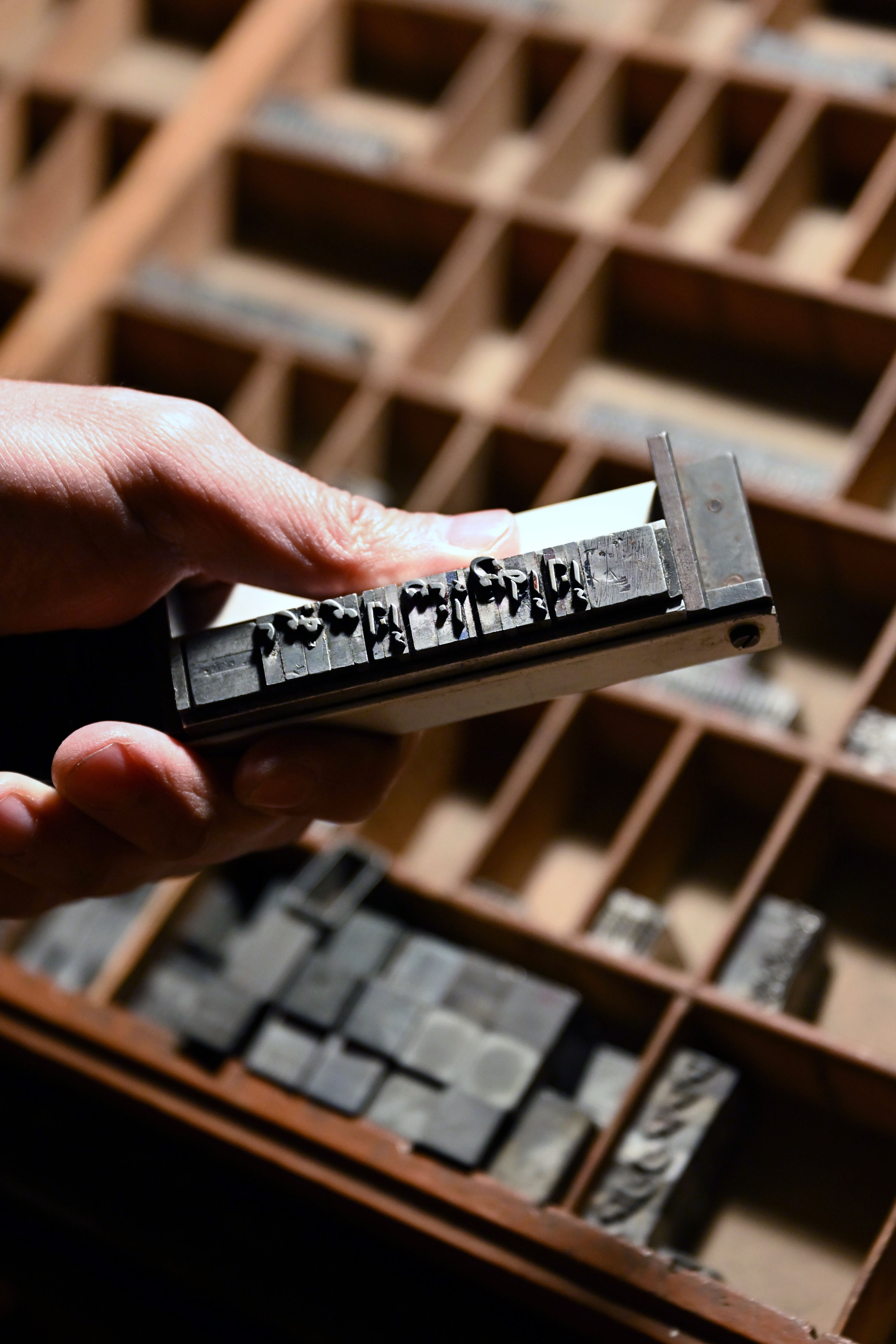 Hand holding a metal composing stick filled with raised movable type above a wooden type case.