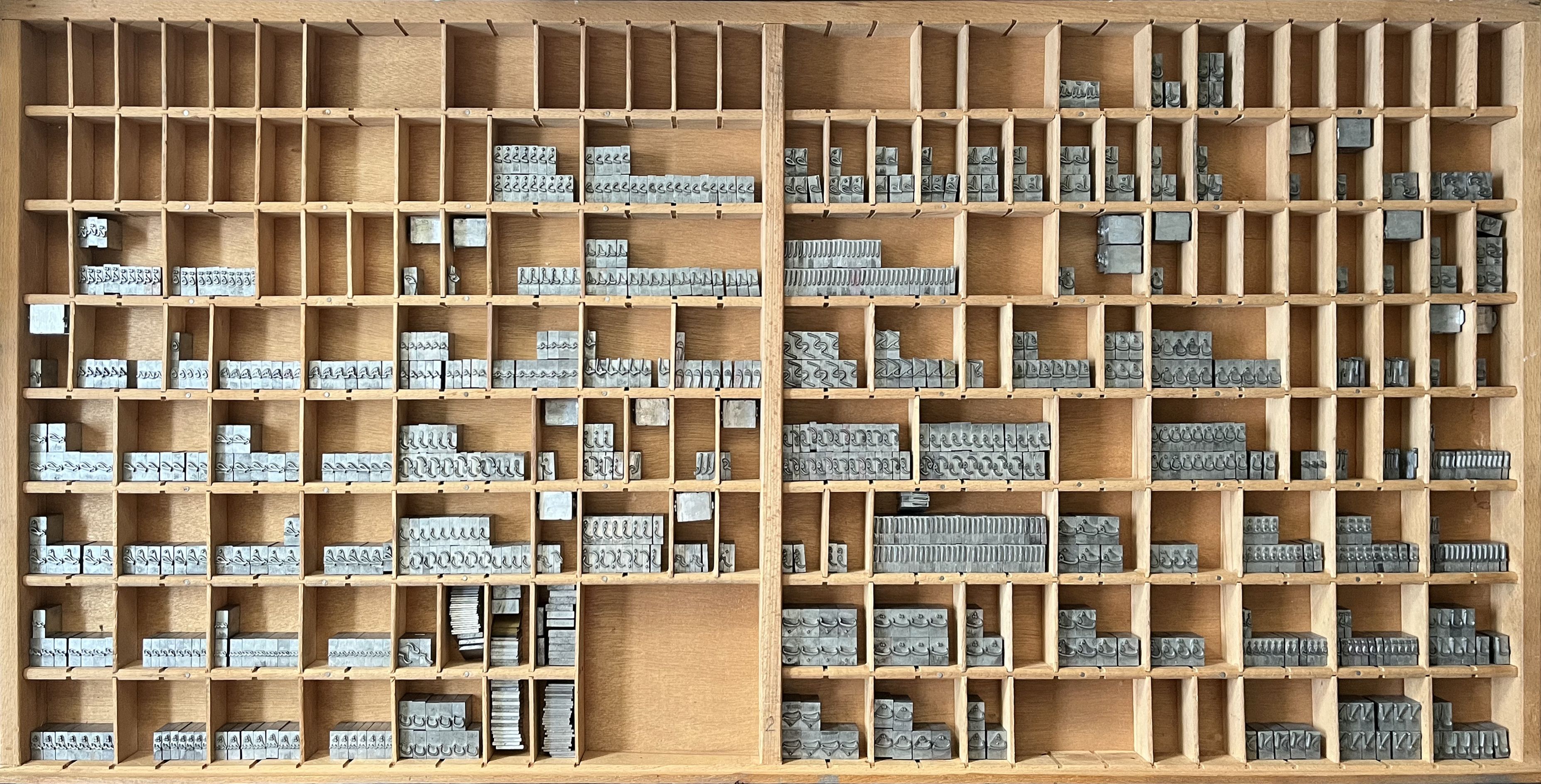 A wooden type case filled with numerous small metal printing blocks, with some compartments left empty.