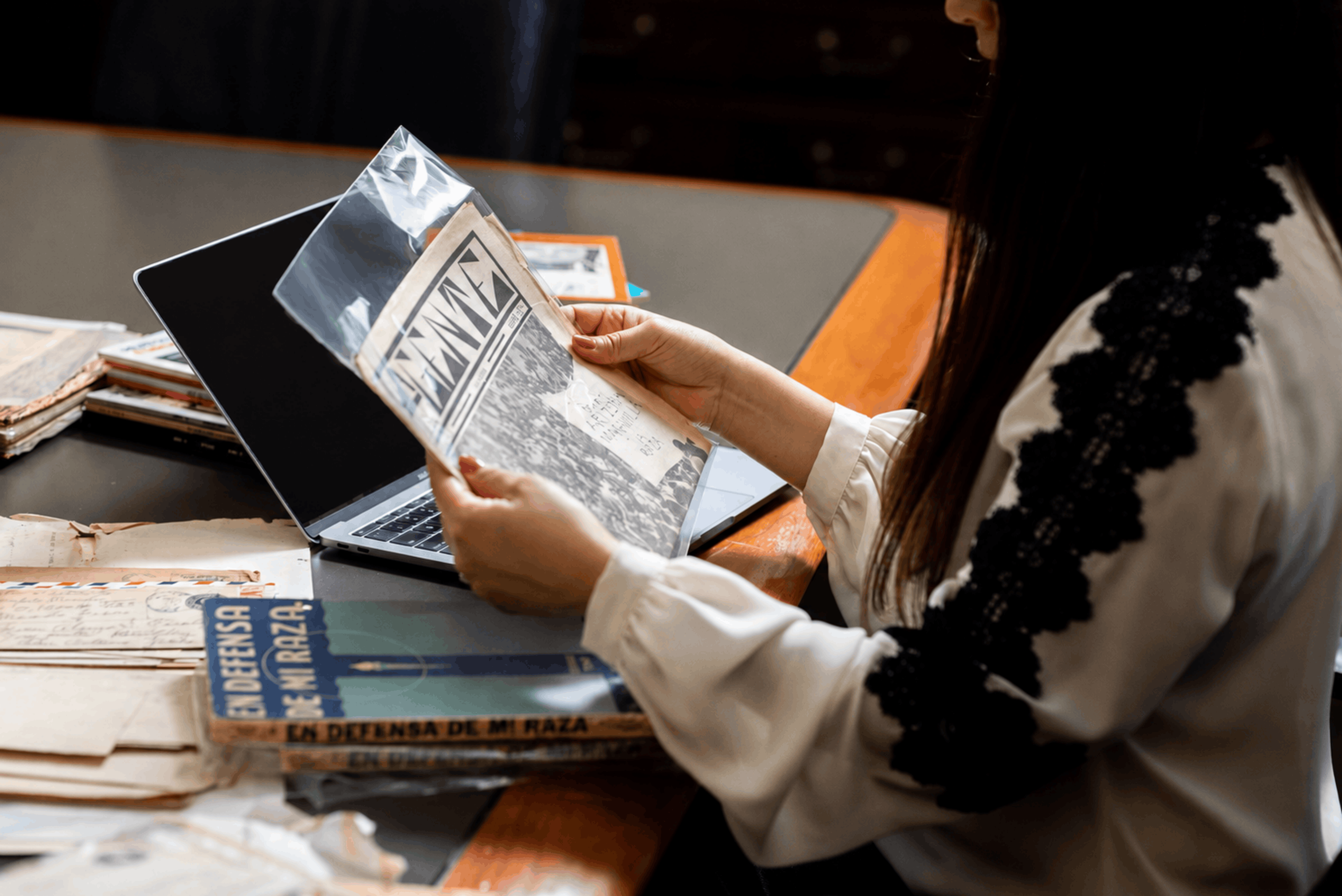 Person reading a black-and-white newspaper at a table beside an open laptop and stacks of printed materials.