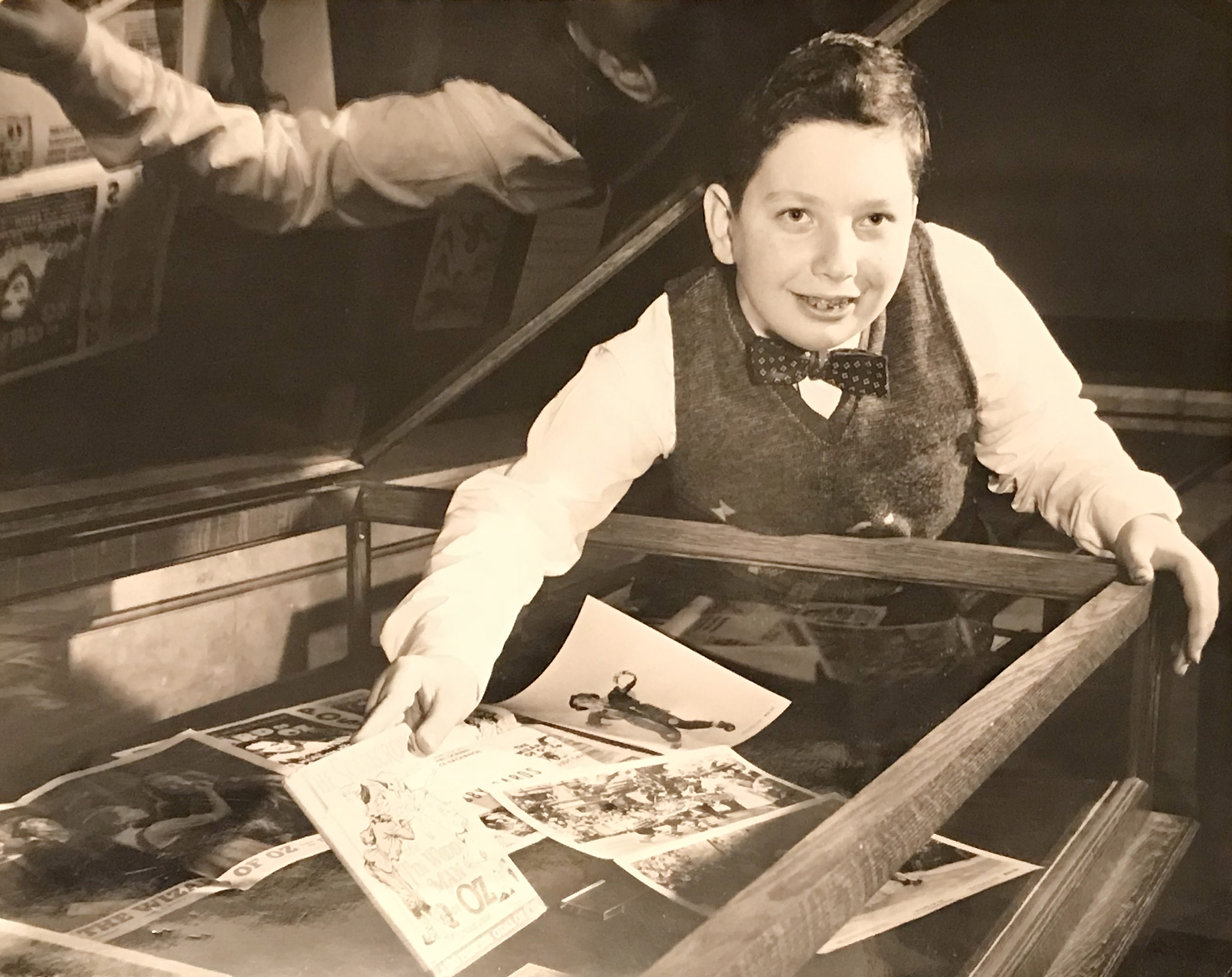 A smiling 12-year-old Justin Schiller in a vest and bow tie leans over a display case with various photographs and printed materials, including a 